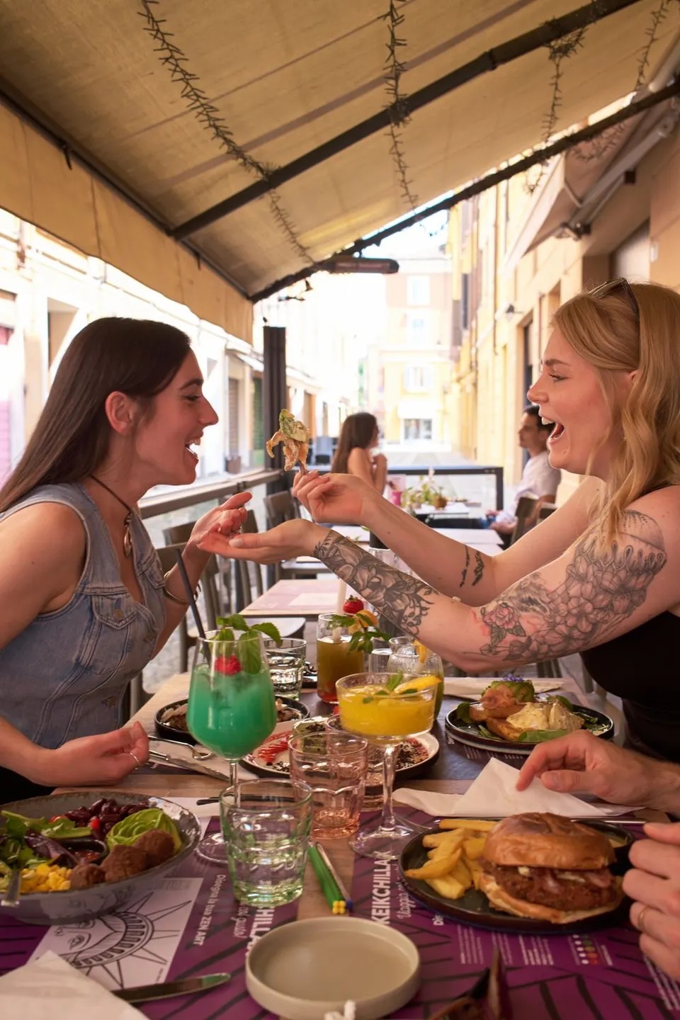 Due amiche si fanno assaggiare i piatti al tavolo esterno di KEIK, tra cocktail colorati, burger e benedict