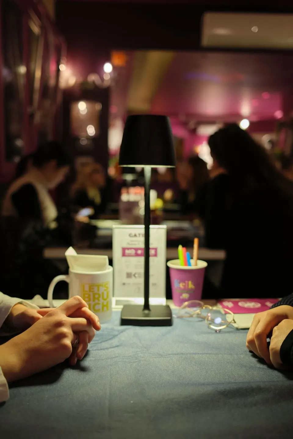 Un tavolo di KEIK la sera, con la lampada accesa, la tazza Let’s Brunch e la sala piena sullo sfondo